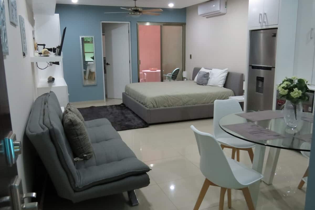 A compact hotel room with a bed, a sofa, a dining table, and a kitchenette.