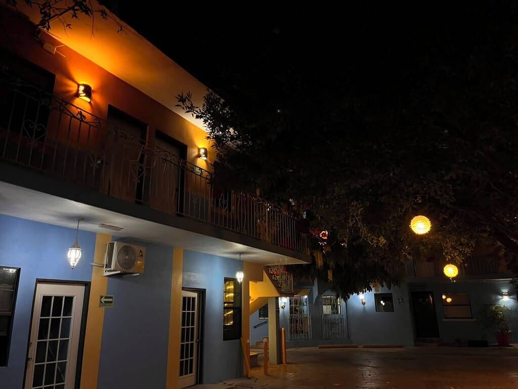 Exterior de un hotel por la noche con balcones iluminados, una escalera y un árbol en primer plano.