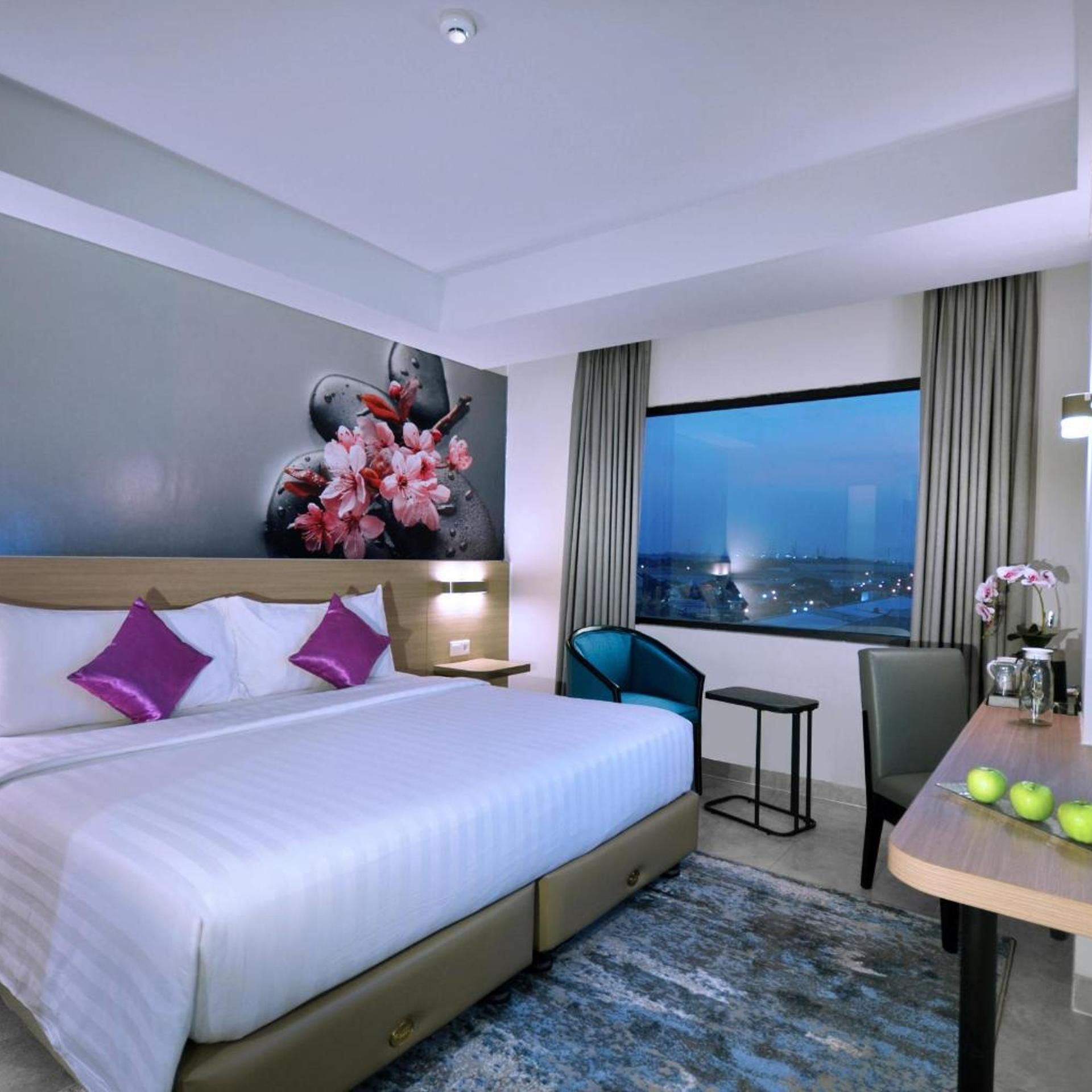 A hotel room with a bed, a desk, a chair, a view of the city at night, and a decorative wall piece.