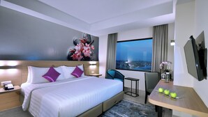 Quarto de hotel moderno com uma cama grande, uma escrivaninha com maçãs, uma cadeira e vista da cidade à noite.