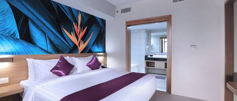 Habitación de hotel con una cama grande, cabecera, mesita de noche, baño visible a través de una puerta abierta y una pared con un cuadro abstracto de gran tamaño.