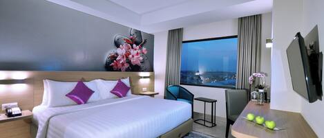 A modern hotel room with a large bed, a desk with apples, a chair, and a view of the city at night.