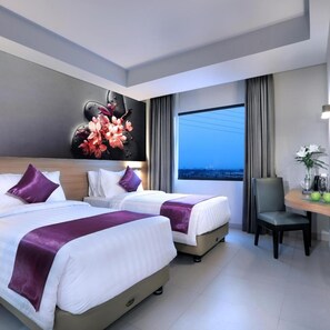 A hotel room with two beds, a desk, a chair, a large window, and a city view.