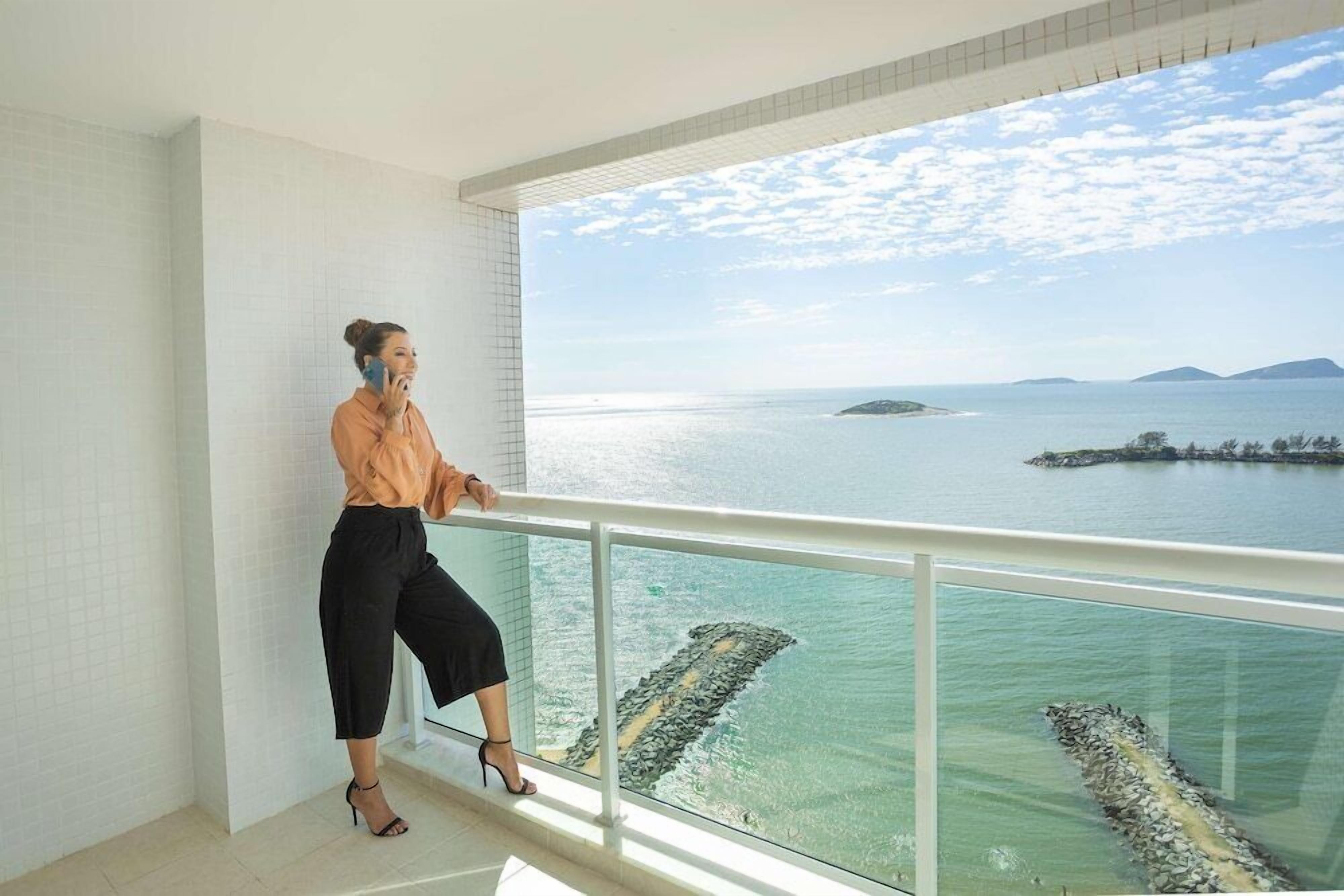 Uma mulher apoiada na grade da varanda, com vista para a costa, céu claro e águas calmas.