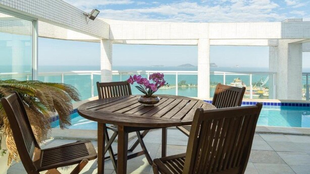 A round wooden table with four chairs and a vase of purple flowers, overlooking a pool and a coastal view. - Golden Tulip Macae (Macaé)
