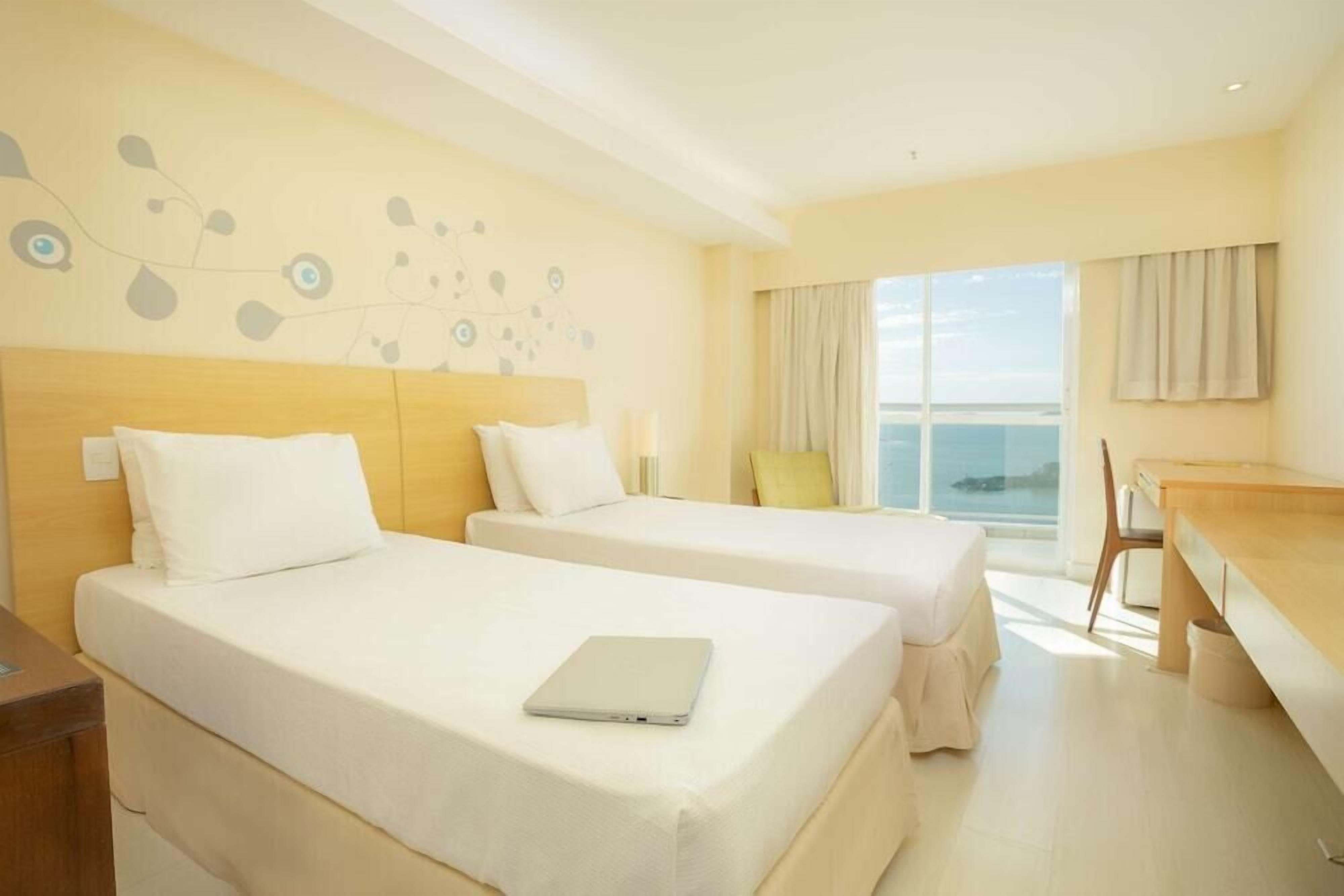 Habitación de hotel con dos camas, una laptop sobre una de ellas, una mesa de comedor y vista al mar.