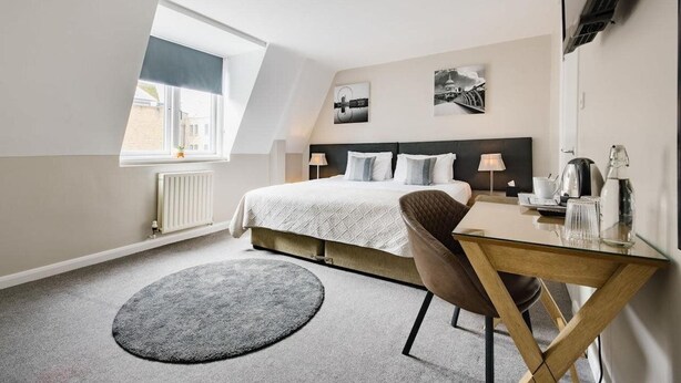 Una habitación de hotel moderna con una cama grande, un escritorio, una silla, un tapete redondo y una ventana con vistas a edificios.