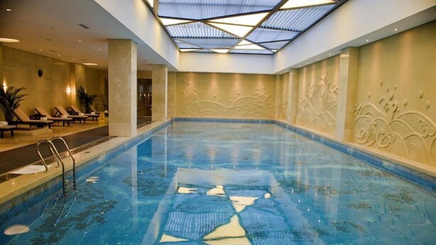 Una piscina interior con techo de cristal, sillones de descanso y paneles decorativos en la pared.