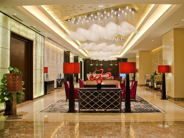 A hotel lobby with a reception desk, red chairs, a red lamp, a chandelier, and a red sculpture.