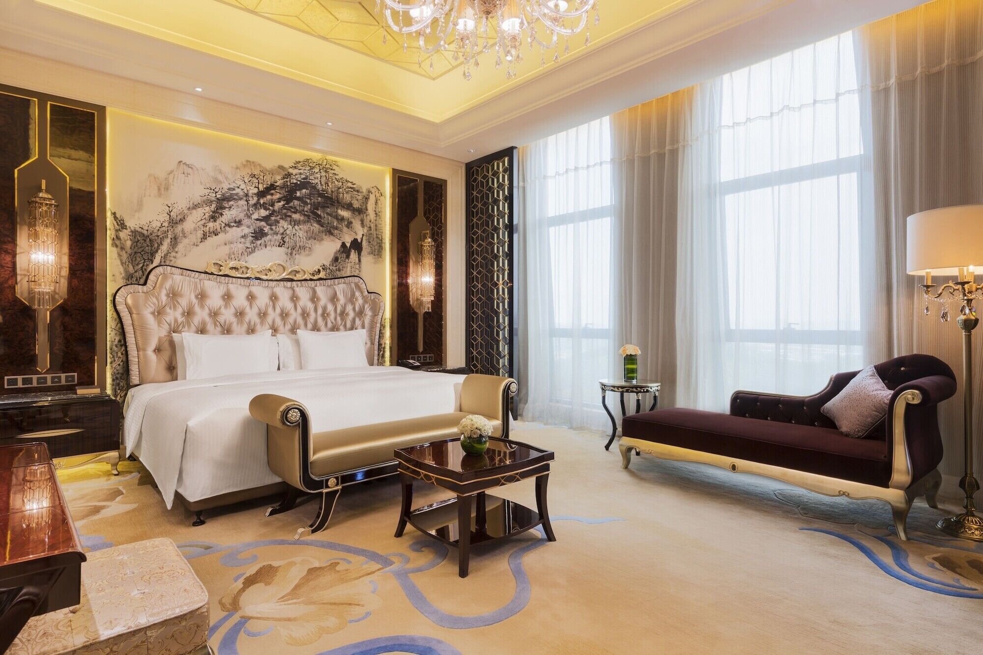 A luxurious bedroom with a large bed, a chaise lounge, a small table, and a chandelier.