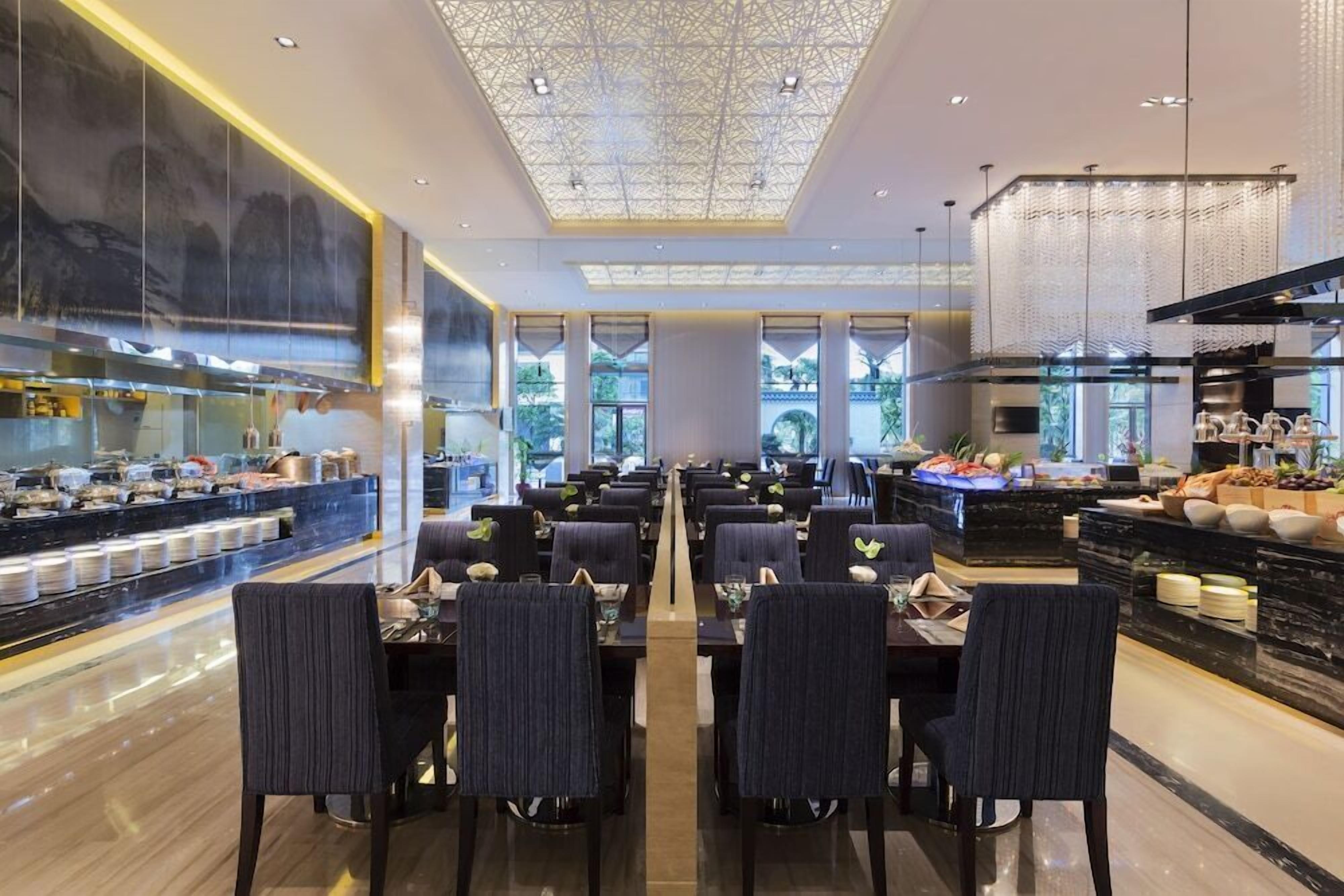 A modern dining area with multiple tables and chairs, a central walkway, and a kitchen area with food displays.