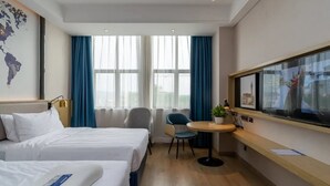 Ett hotellrum med två sängar, ett skrivbord, en stol, ett litet bord med en växt och ett stort fönster med gardiner.