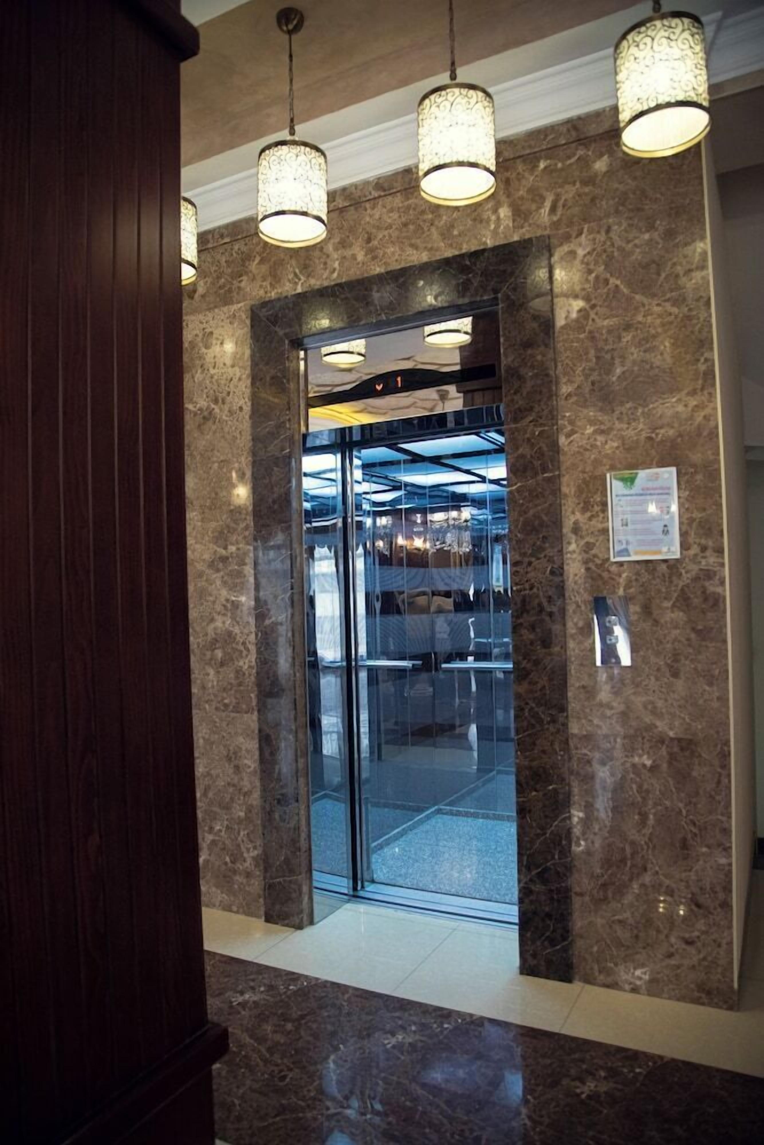 A marble wall with a glass elevator entrance, a wooden panel to the left, and a hanging light fixture above.