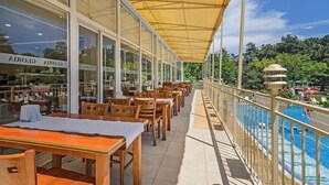 A covered outdoor seating area with tables and chairs, overlooking a pool and greenery. - Gloria Saints Constantine And Helena (Varna)
