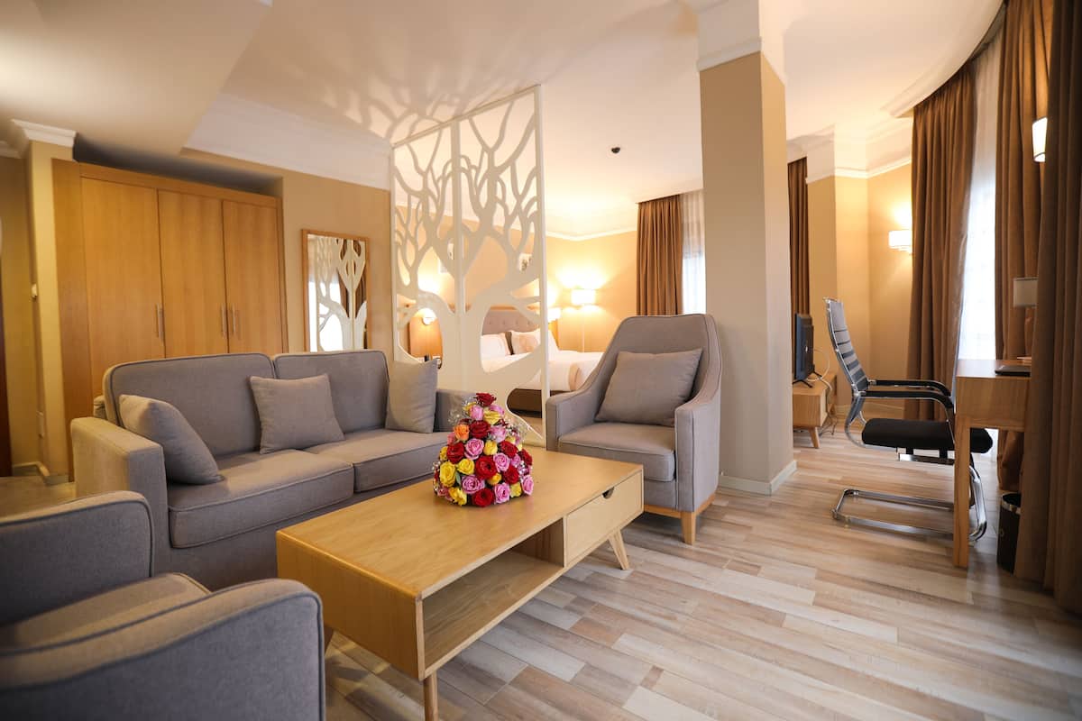A modern hotel room with a sofa set, a coffee table with a floral arrangement, a desk with a chair, and a decorative screen with a tree design.