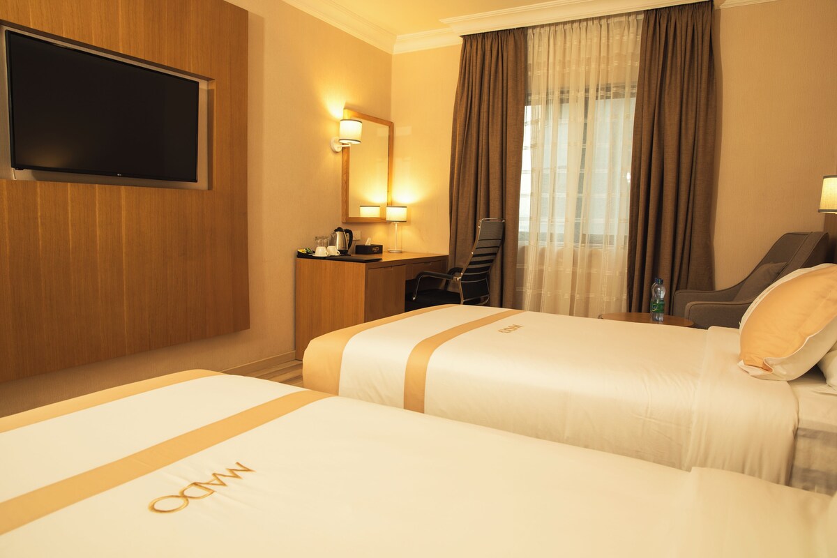 A hotel room with two beds, a TV, a desk with a chair, a mirror, and a window with curtains.