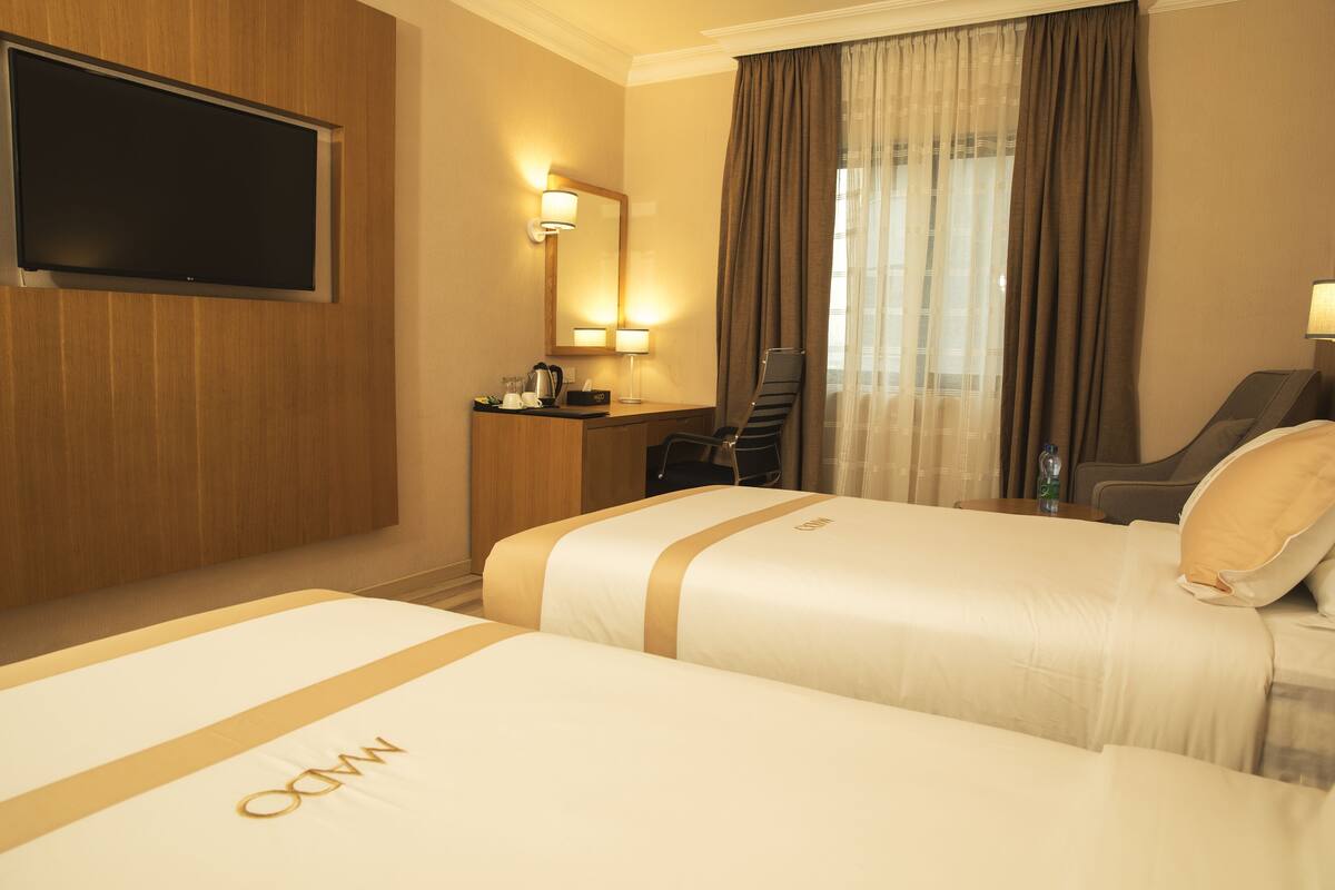 A hotel room with two beds, a TV mounted on the wall, a desk with a chair, a mirror, and a window with curtains.
