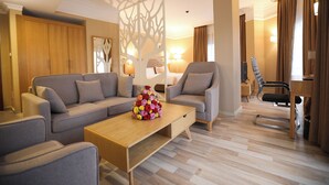 Una habitación de hotel moderna con sofá, sillones, una mesa de centro de madera con un centro de mesa floral y un escritorio con una silla.
