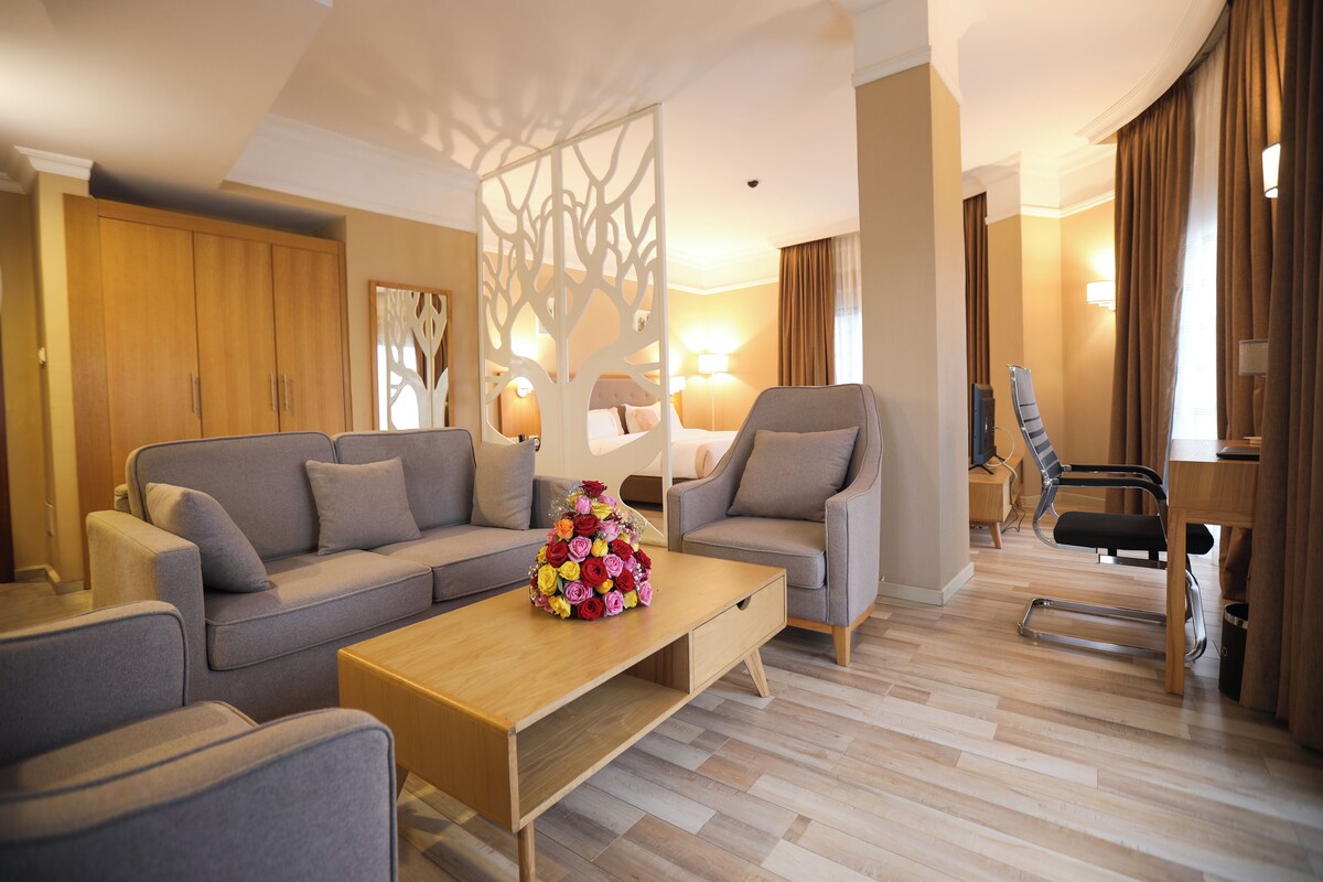 A modern hotel room with a sofa, armchairs, a wooden coffee table with a floral centerpiece, and a desk with a chair.