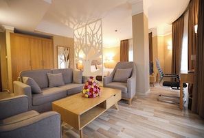 A modern hotel room with a sofa, armchairs, a wooden coffee table with a floral centerpiece, and a desk with a chair. - Mado Hotel (Addis Ababa)
