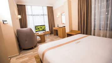 A hotel room with a large bed, a desk with a computer, a chair, a small table, and a window with curtains.