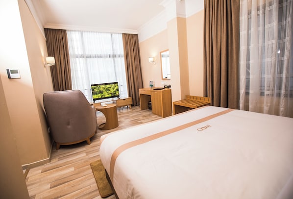 Hotelkamer met een groot bed, een bureau met een computer, een stoel, een klein tafeltje en een raam met gordijnen.