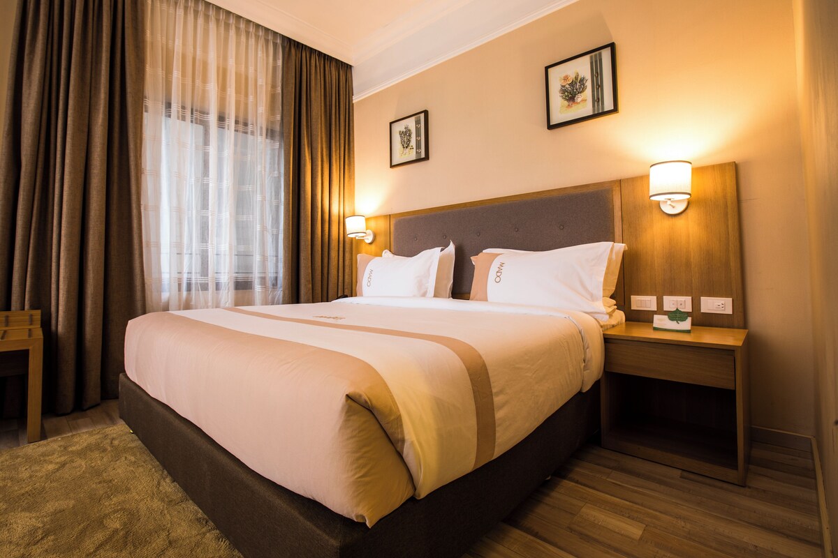 Habitación de hotel con una cama grande, dos mesitas de noche, un tocador de madera, un banco de madera y un amplio ventanal con cortinas.