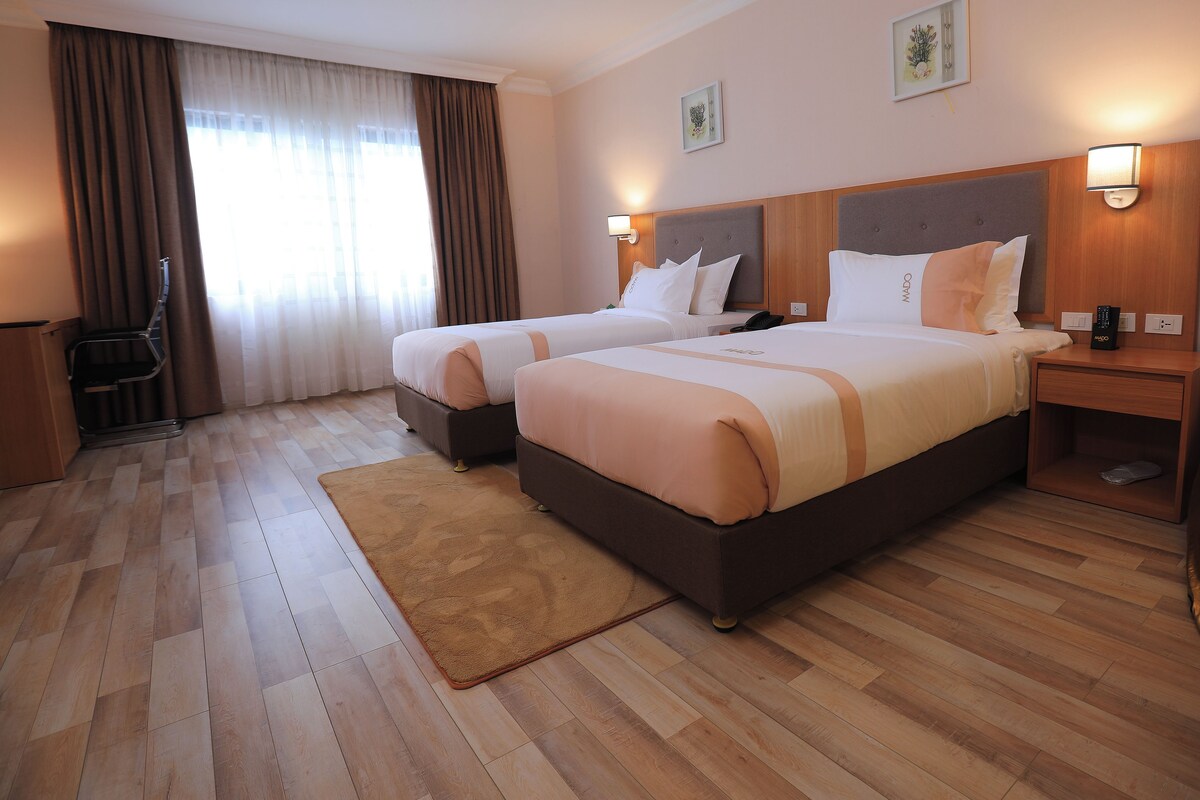 A hotel room with two beds, a wooden floor, a desk, a chair, and a window with curtains.