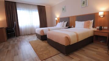 A hotel room with two beds, a wooden floor, a desk, a chair, and a window with curtains.