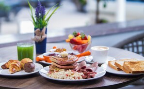 Una mesa con un desayuno que incluye cruasanes, huevos revueltos, salchichas, tostadas y un bol de fruta.