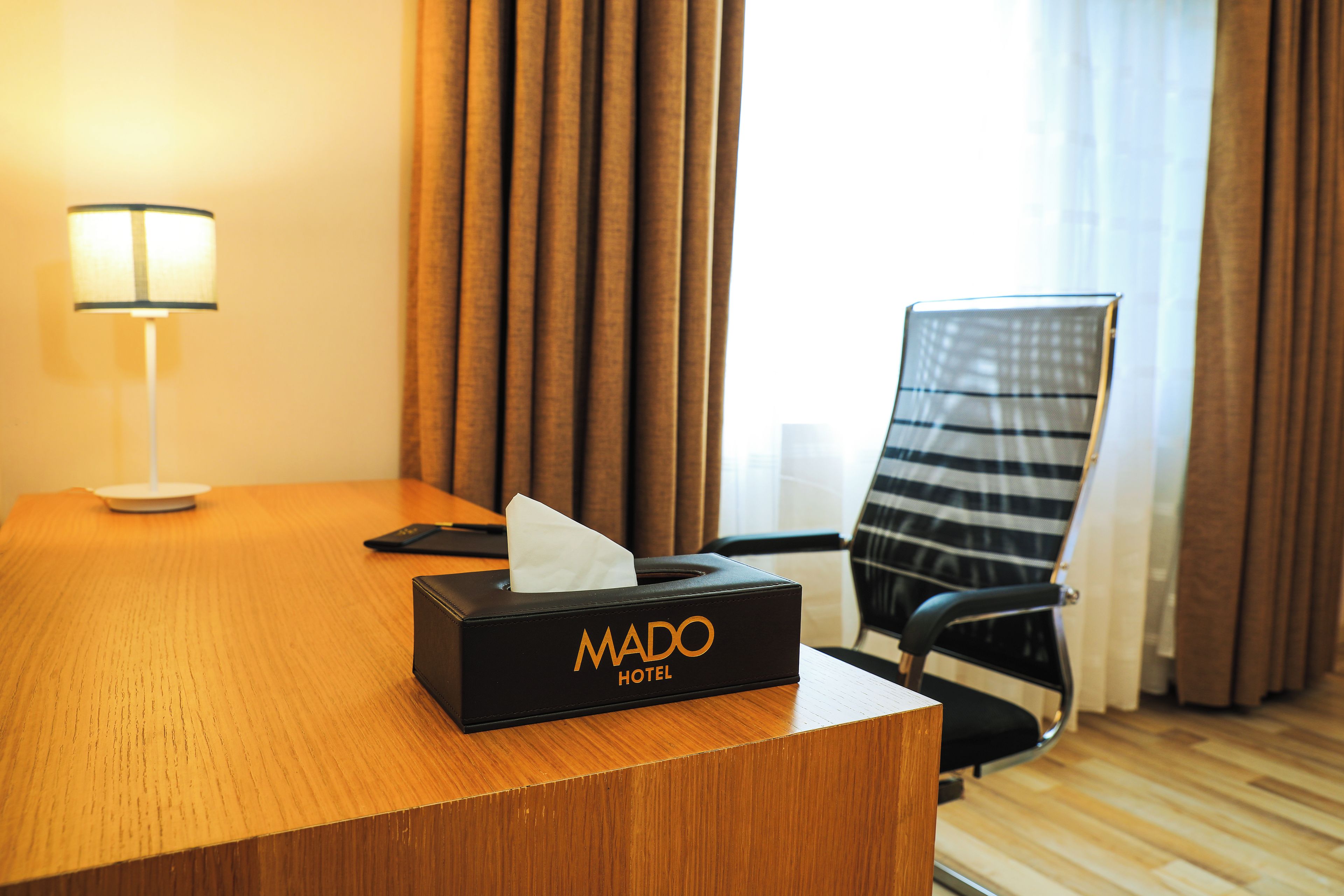 Hotellihuone, jossa on puinen työpöytä, musta paperipyyhekotelo, jonka kyljessä lukee ”MADO HOTEL”, musta toimistotuoli ja lamppu.