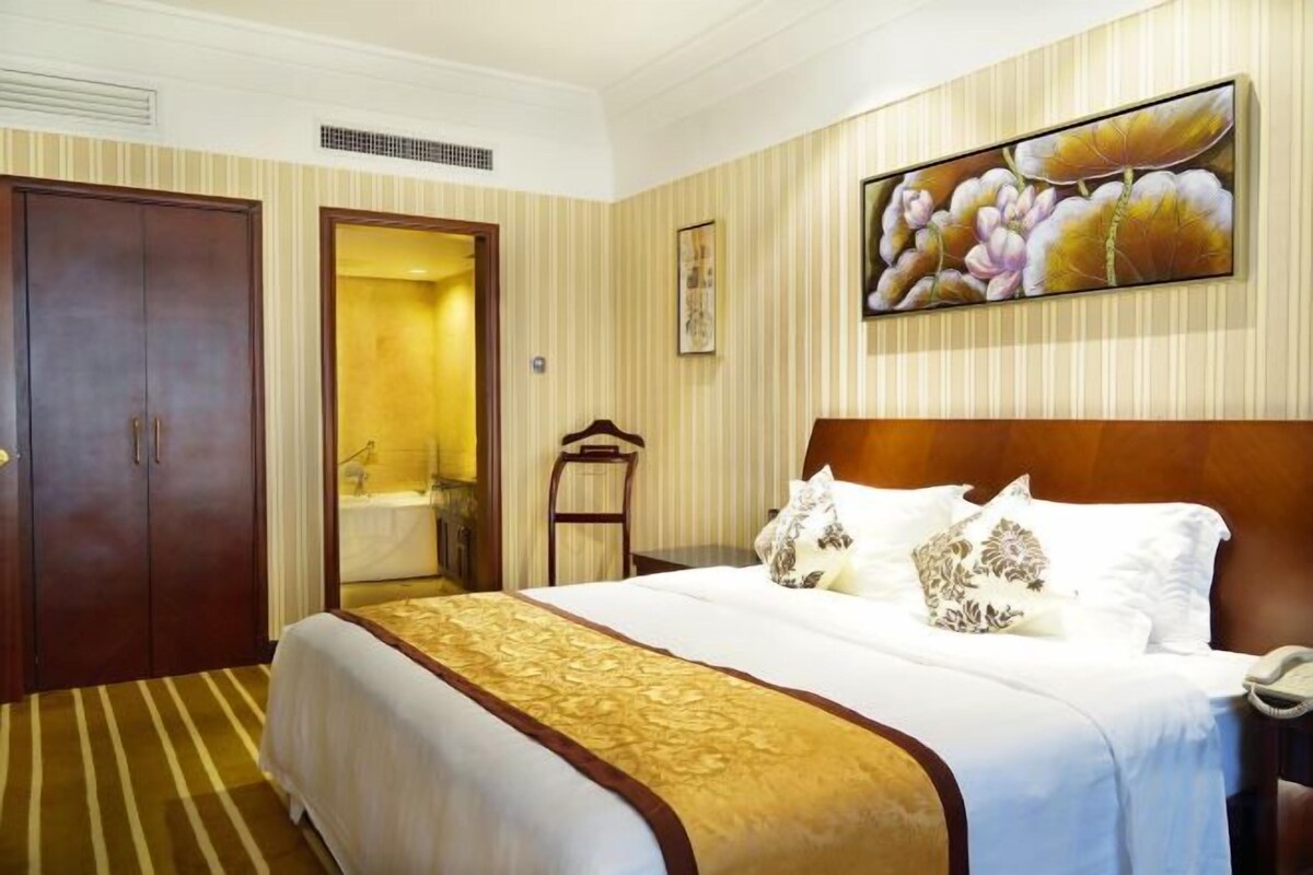 A hotel room with a large bed, a wooden headboard, a painting above the bed, and a door leading to a bathroom.