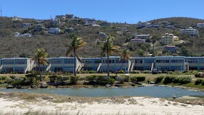 A coastal area with a beach, a small body of water, and a row of blue buildings with a hill in the background.