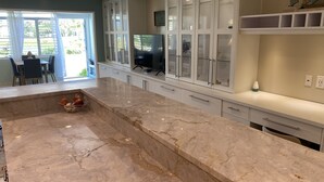 A modern kitchen with a marble countertop, white cabinetry, and a glass-enclosed dining area.