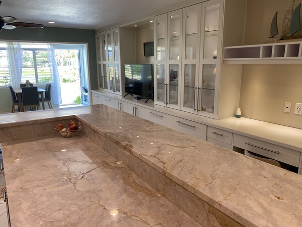 A modern kitchen with a marble countertop, white cabinetry, and a glass-enclosed dining area.