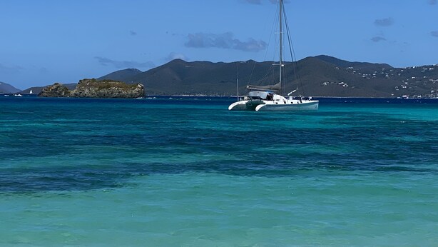 A beach with clear turquoise water, a sailboat anchored near the shore, and a mountainous backdrop.