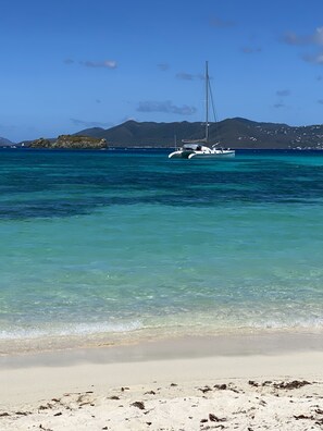 A beach with clear turquoise water, a sailboat anchored near the shore, and a mountainous backdrop.