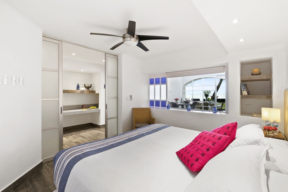 Una habitación de hotel moderna con cama, ventilador de techo, armario, una mesita de noche con lámpara y ventana con vistas al exterior.