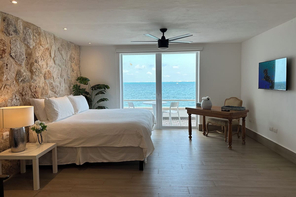 Un dormitorio con una cama, una mesita de noche, una mesa y una silla. Hay un ventanal con vista al océano.