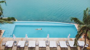 Piscina infinita com uma pessoa nadando, rodeada por cadeiras de praia e guarda-sóis, com vista para um mar azul e límpido.