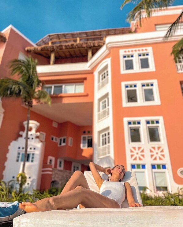 Uma pessoa relaxando numa cadeira de lounge branca perto de uma piscina, com um edifício com varandas e janelas decorativas ao fundo.