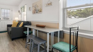 A cozy living room with a gray sofa, a wooden side table, and a window with blinds. - Gypsea Wind: Oceanside, Short Walk to Beach, Short Stays Anytime! (Kill Devil Hills)