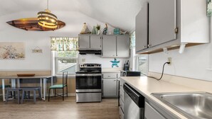 A modern kitchen with stainless steel appliances, a dining area with a table and chairs, and decorative items on the cabinets. - Gypsea Wind: Oceanside, Short Walk to Beach, Short Stays Anytime! (Kill Devil Hills)