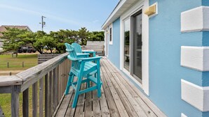 A wooden deck with two turquoise Adirondack chairs, a blue building with white trim, and a garage in the background. - Gypsea Wind: Oceanside, Short Walk to Beach, Short Stays Anytime! (Kill Devil Hills)