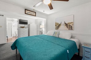 A bedroom with a teal bedspread, a ceiling fan, a beach-themed artwork, and a dresser with a mirror.