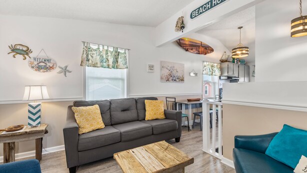 A cozy living room with a gray sofa, wooden coffee table, and blue armchair. A kitchen area is visible in the background with a dining table and chairs. - Gypsea Wind: Oceanside, Short Walk to Beach, Short Stays Anytime! (Kill Devil Hills)