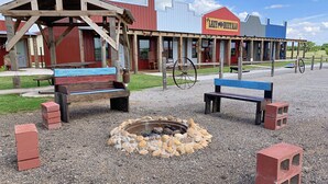 Un espacio rústico al aire libre con bancos y una chimenea, un edificio rojo con el rótulo "The Lazy Buffalo", y una construcción azul en el fondo.