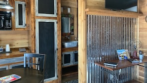 Interior rústico de una cabaña con paredes de madera, una pizarra, microondas, lavabo, mesa con sillas y televisor.