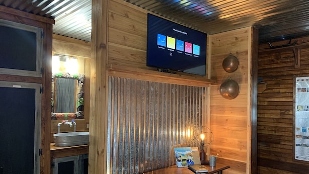 Un acogedor interior de cabaña con pared de madera, un televisor montado que muestra opciones de Netflix, una mesita con un libro y una lámpara, y una decoración rústica.