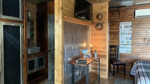 Una habitación acogedora con techo de madera, una cama, una mesa con un libro y una lámpara, una silla y un tablón de anuncios con información.